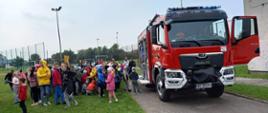 Przed budynkiem szkoły podstawowej stoi samochód ratowniczo- gaśniczy z otwartymi skrytkami i drzwiami Przed samochodem stoją uczniowie wraz z opiekunami i strażacy prezentujący sprzęt strażacki. W tle boisko szkolne.