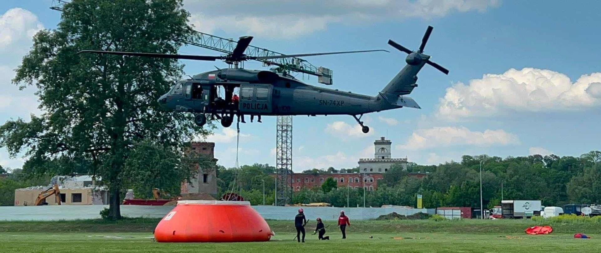 Śmigłowiec Black Hawk zawisa nad trawiastym placem ćwiczeń podczas operacji z użyciem zbiornika Bambi Bucket. Na ziemi strażacy przygotowują sprzęt do podjęcia ładunku.