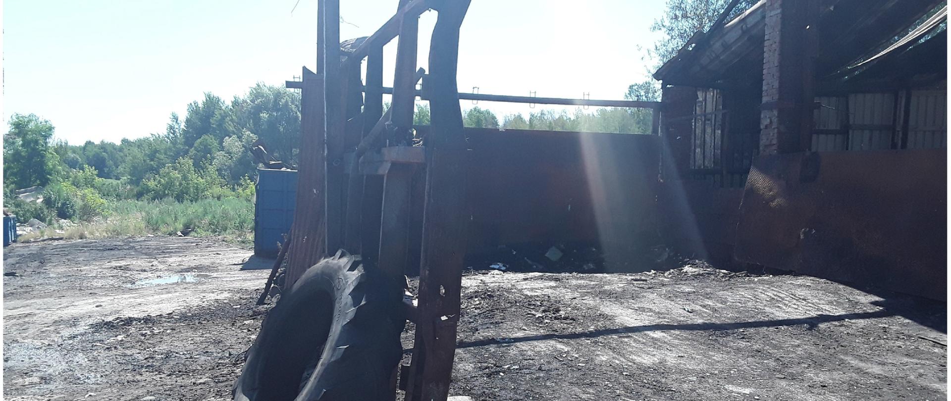 Pokryte czarnym nalotem uprzątnięte pogorzelisko: po prawej stronie widać parterowy, osmalony budynek, w środku stalowa kratownica, pokryta sadzą i oparta o nią opona od ciągnika. W tle widoczne zielone drzewa.