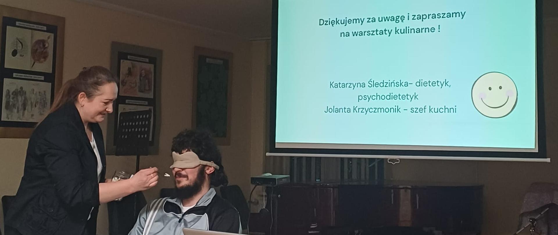 Na zdjęciu w sali konferencyjnej - auli wrocławskiej bursy widać młodego chłopca siedzącego za stołem. ma on na oczach opaskę zasłaniającą widzenie. Obok niego stoi pani, która podaje mu łyżeczką do ust potrawę do spróbowania. W tle widać włączony ekran z prezentacja jednego ze slajdów mówiących o zdrowym odżywianiu.