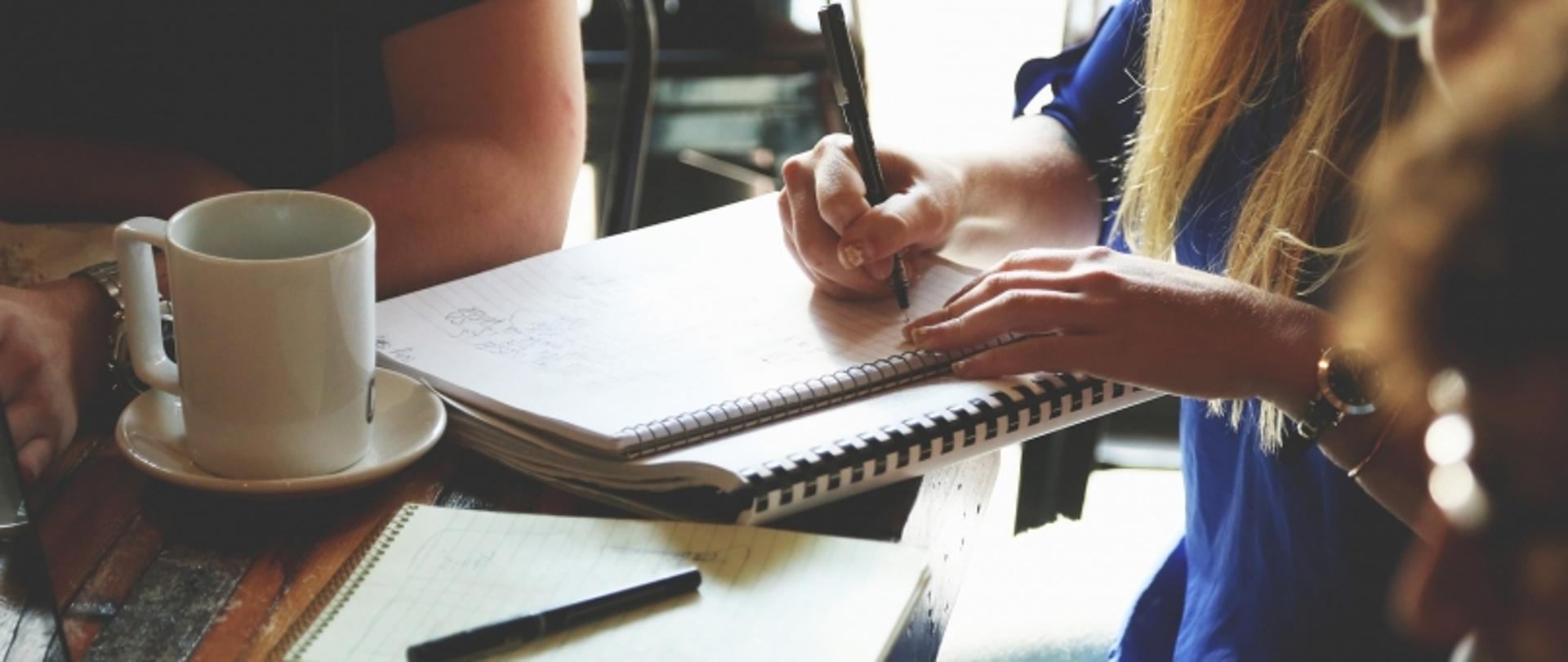 „Grupa osób siedząca przy stole, pisząca w zeszytach i notatnikach podczas nieformalnego spotkania.”