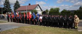 Strażacy ze sztandarami stoją w szyku przed trawnikiem. Za nimi stoi dom z brązowym dachem. 