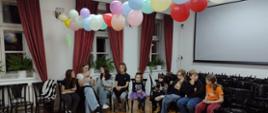 Na zdjęciu widnieje grupa dzieci, którzy siedzą na krzesłach w sali koncertowej. Sala jest udekorowana kolorowymi balonami