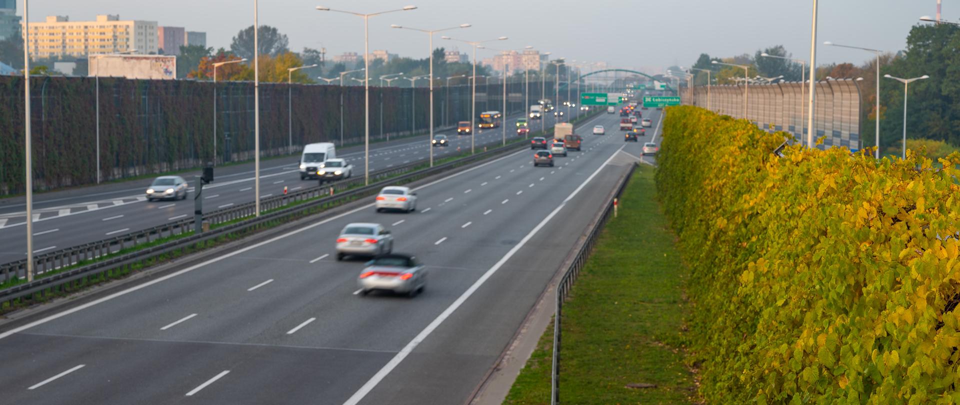 S8_Trasa_Torunska, ciąg główny trasy. Widać poruszające sie pojazdy. Po prawej stronie widać porośnięty zielonym pnączem ekran akustyczny.