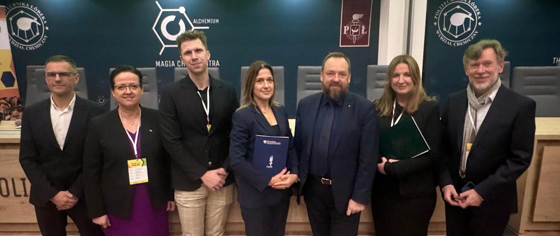 Grupowe zdjęcie siedmiu osób stojących w eleganckiej sali konferencyjnej. Ludzie mają na sobie garnitury i eleganckie stroje. Dwie kobiety trzymają w dłoniach teczki lub foldery. Za nimi znajduje się ciemnozielona lub granatowa ściana z dwoma dużymi znakami graficznymi uczelni oraz herbem. W tle widać rząd pustych krzeseł i elementy drewnianej ławy. Panuje oficjalna atmosfera, sugerująca wydarzenie akademickie lub naukowe.