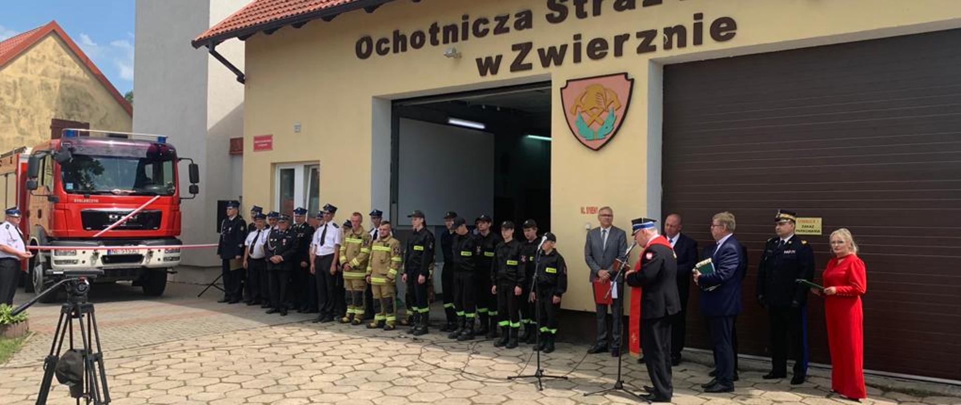 Po lewej stronie znajduje się przekazany pojazd pożarniczy marki MAN, obok po prawej stronie na tle remizy ochotniczej straży pożarnej w Zwierznie stoją druhowie oraz zaproszeni goście.