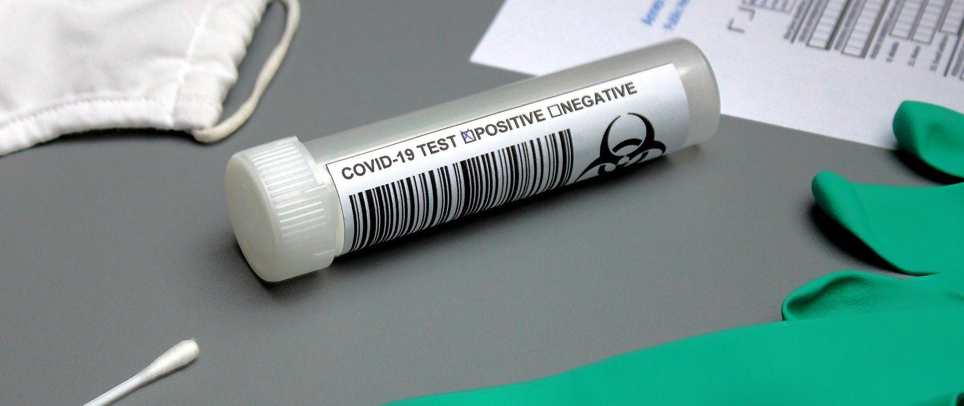 na stole leżą patyczki z próbek do pobierania testów na koronawirusa, rękawiczki jednorazowe, maseczka ochronna oraz formularz, a na środku leży probówka z oznaczeniem Covid19 test positive 