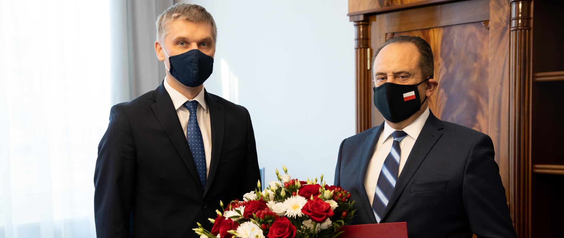 Obok siebie stoją Piotr Nowak i Andrzej Gut-Mostowy, który trzyma w ręku teczkę i bukiet biało-czerwonych kwiatów.