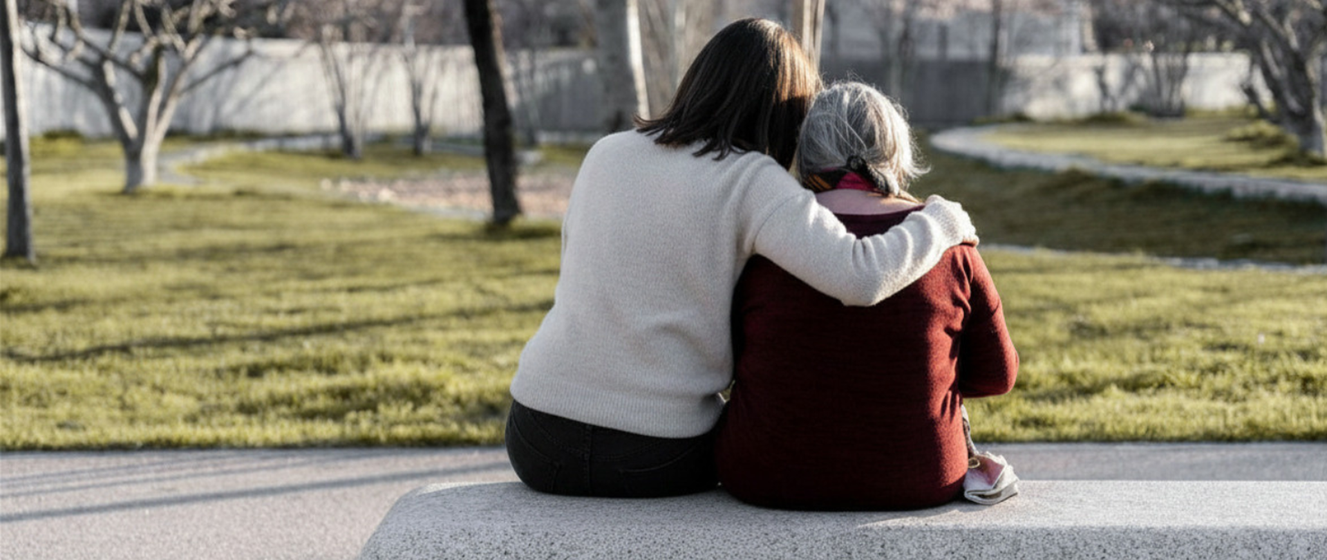 dwie kobiety, młodsza i starsza siedzą na ławce, młodsza obejmuje ramieniem seniorkę