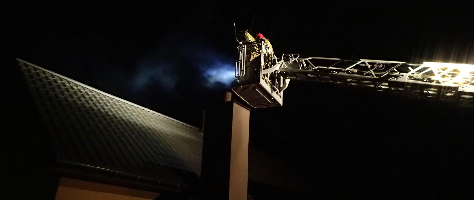 2 ratowników w koszu drabiny mechanicznej gaszących pożar sadzy w przewodzie kominowym, pora nocna