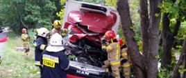 strażacy stoją przy uszkodzonym rozbitym po uderzeniu w drzewo samochodu dostawczego koloru czerwonego 