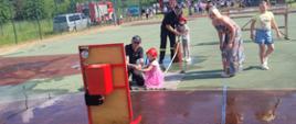 Zdjęcie obrazuje strażaków z OSP Sorbin, którzy przeprowadzają konkurs dla dzieci podczas festynu w Sorbinie. Prowadzona konkurencja odbywa się na boisku szkolnym. Dzieci z hydronetki strażackiej leją wodę do nalewaka.