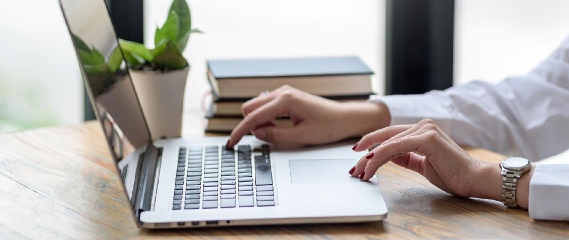 Dłonie kobiety na klawiaturze laptopa, który leży na biurku. Obok laptopa notatniki i długopis.