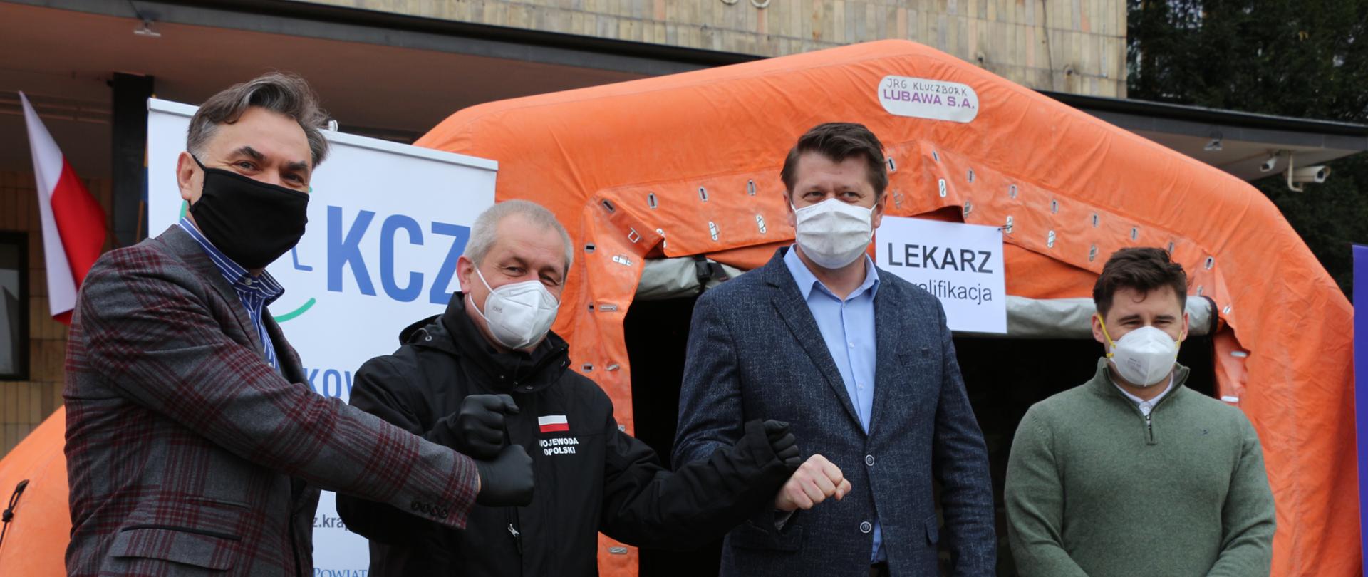 Na zdjęciu 4 mężczyzn, stojących na tle pomarańczowego namiotu polowego. Mężczyźni mają ułożne dłonie w piąstki, którymi się uderzają na znak sukcesu. Patrzą w obiektyw. 