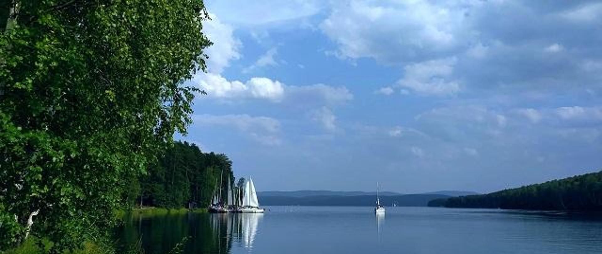żaglówka pływająca po jeziorze, kilka żaglówek przycumowanych do brzegu z widokiem na las 