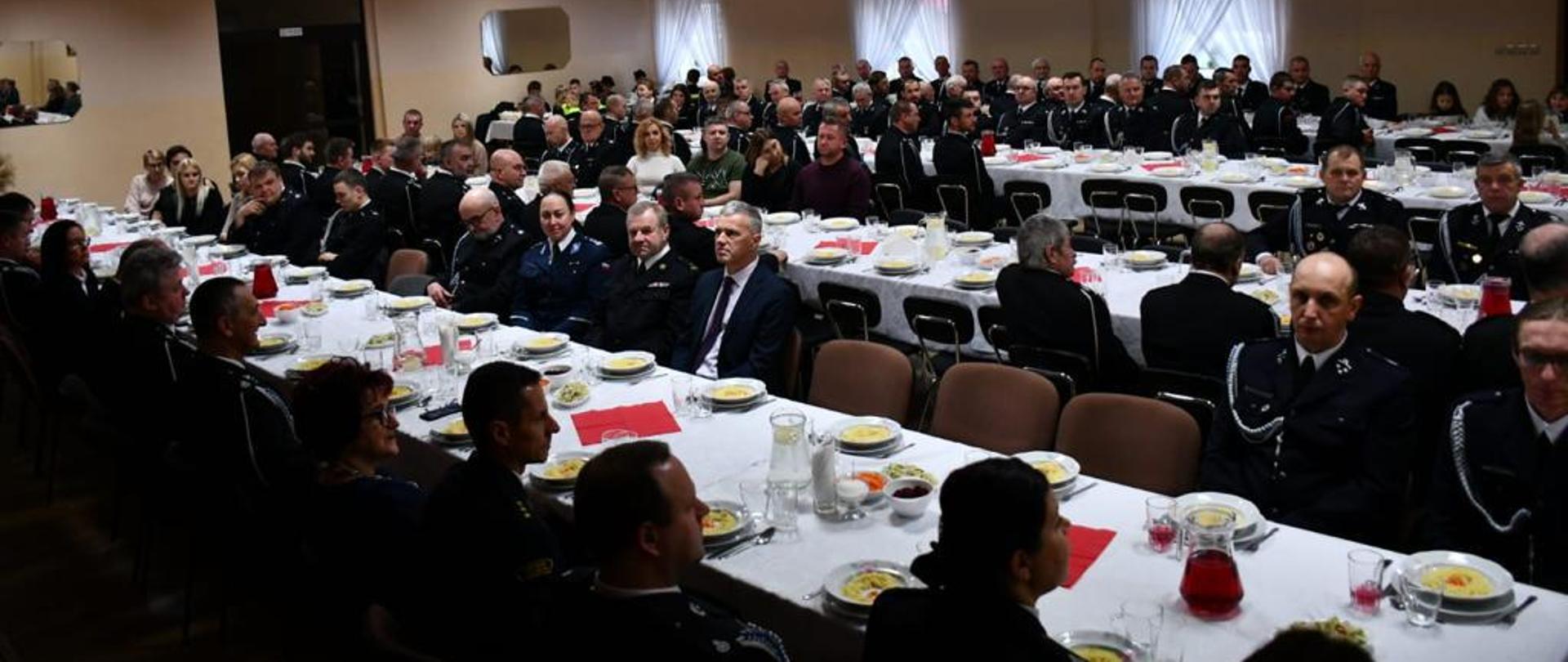 Zdjęcie wykonane wewnątrz sali wiejskiej. Zdjęcie przedstawia strażaków z powiatu wolsztyńskiego zasiadających przy stole. Strażacy ubrani w mundury wyjściowe.