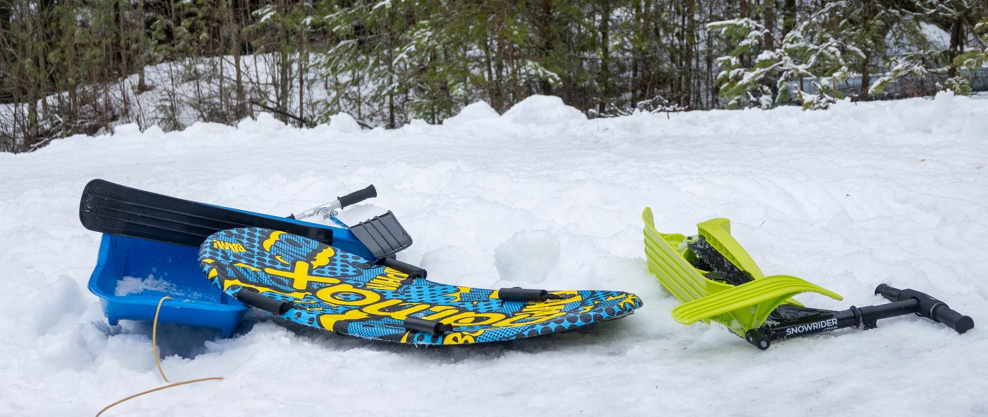 w tle las, a na pierwszym planie na śniegu pozostawione sanki, pługi i deska do zjeżdżania