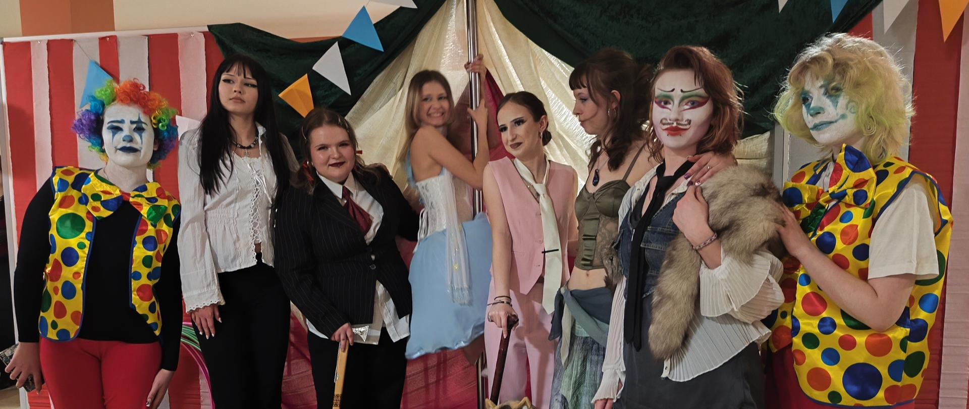 Zdjęcie przedstawia grupę osób przebranych w stroje inspirowane cyrkiem, ustawionych wspólnie przed dekoracją przypominającą namiot cyrkowy. W tle widoczne są kolorowe proporczyki, balony oraz fragment czerwono‑białej scenografii kojarzącej się z wejściem do cyrku.