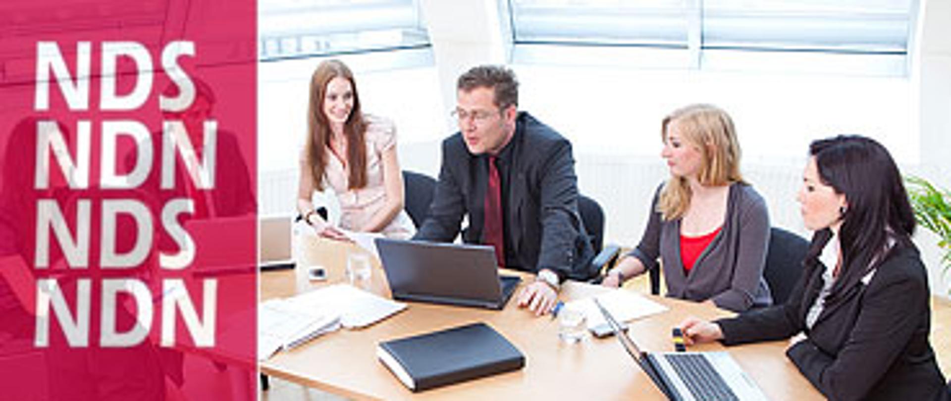 na zdjęciu widnieją rozmawiający ludzie z laptopami przy biurkach 