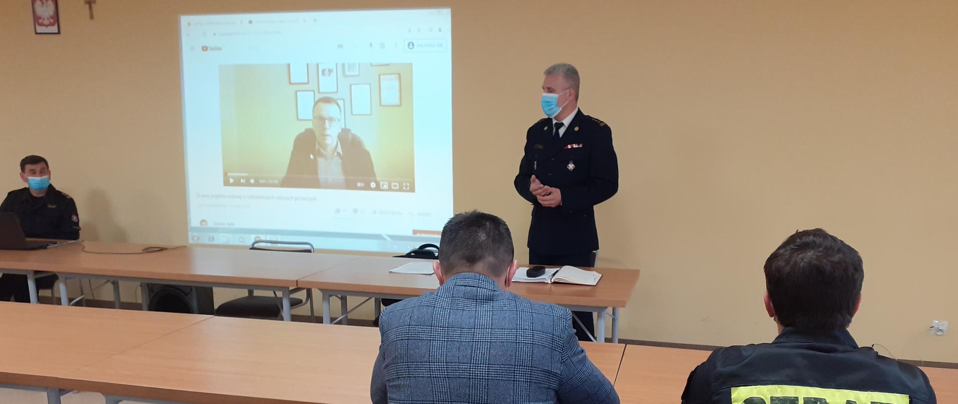 Na zdjęciu widzimy salę w której zebrali się przedstawiciele Ochotniczych Straży Pożarnych, samorządów oraz Komendy Powiatowej PSP. Na ścianie przedstawiony jest materiał video, w którym prawnik przedstawia założenia projektu ustawy o OSP. Stoi Komendant Powiatowy PSP przekazując informacje zaproszonym przedstawicielom z gmin powiatu myślenickiego.