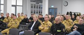 Funkcjonariusze Państwowej Straży Pożarnej oraz osoby cywilne siedzą na krzesłach obok siebie i słuchają strażacy są w umundurowaniu koloru czarnego i musztardowego. 