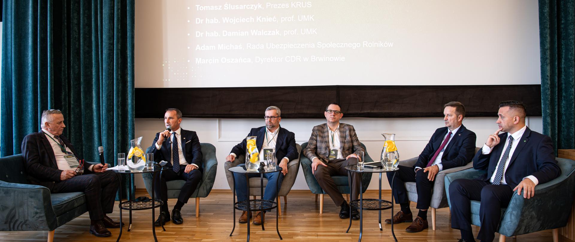 Na scenie sześciu mężczyzn siedzi w fotelach podczas panelu dyskusyjnego II Konferencji Naukowej KRUS - strategia, zdrowie i bezpieczeństwo polskiej wsi. W tle widoczny tytuł konferencji oraz imiona, nazwiska oraz stanowiska panelistów.