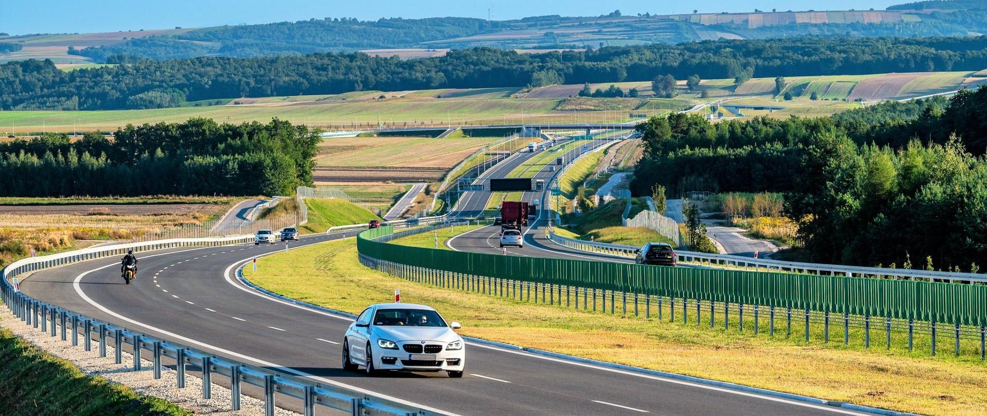 Droga ekspresowa S7 na odcinku Moczydło - Miechów. Dwujezdniowa droga ekspresowa wijąca się między polami i lasami po niewielkich pagórkach. Widoczne pojazdy poruszające się w obu kierunkach.