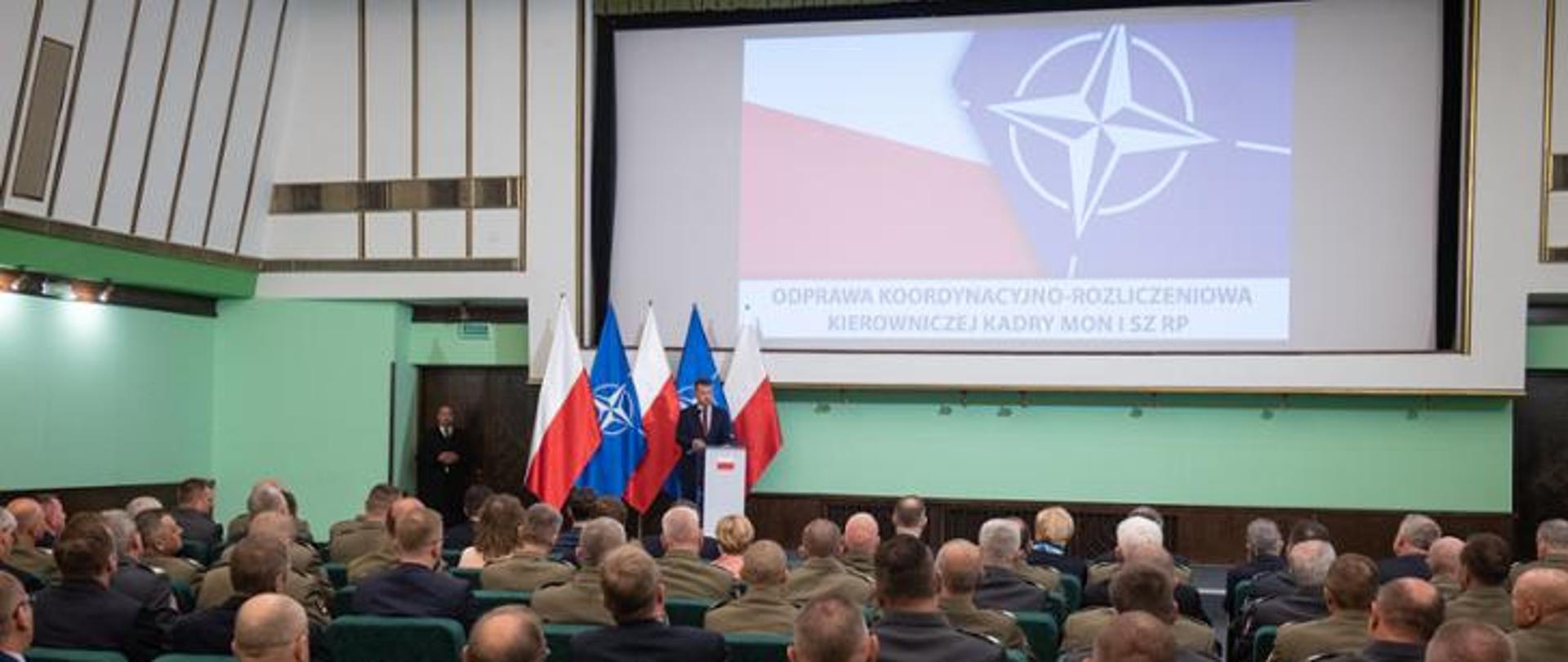 Jeden mężczyzna stojący przy mównicy i wypowiadający się do wielu siedzących osób na widowni