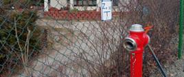 Zdjęcie przedstawia przeciwpożarowy hydrant zewnętrzny w kolorze czerwonym, który jest na ogrodzeniu oznakowany.