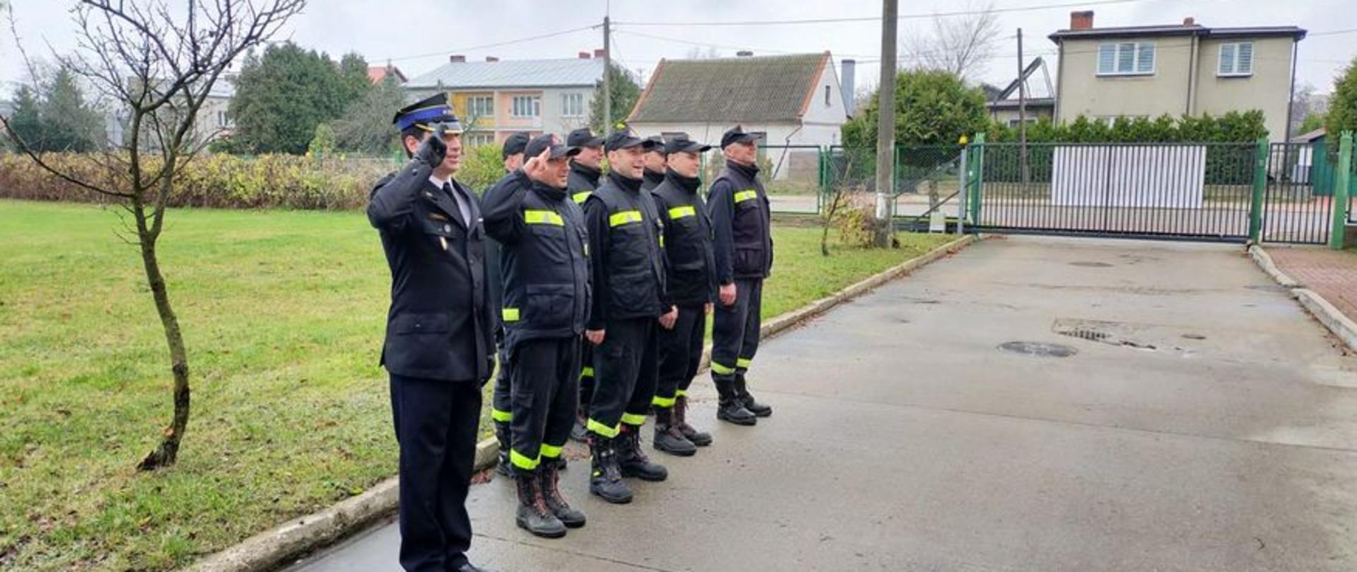 Na drodze przy zielonym trawniku stoi w dwuszeregu ośmiu strażaków w czarnych mundurach. Dwóch z nich salutuje.