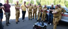 Strażacy w mundurach koloru musztardowego oraz osoby cywilne stoją w kręgu obok siebie w środku jest skrzynia ze sprzętem informatycznym. 