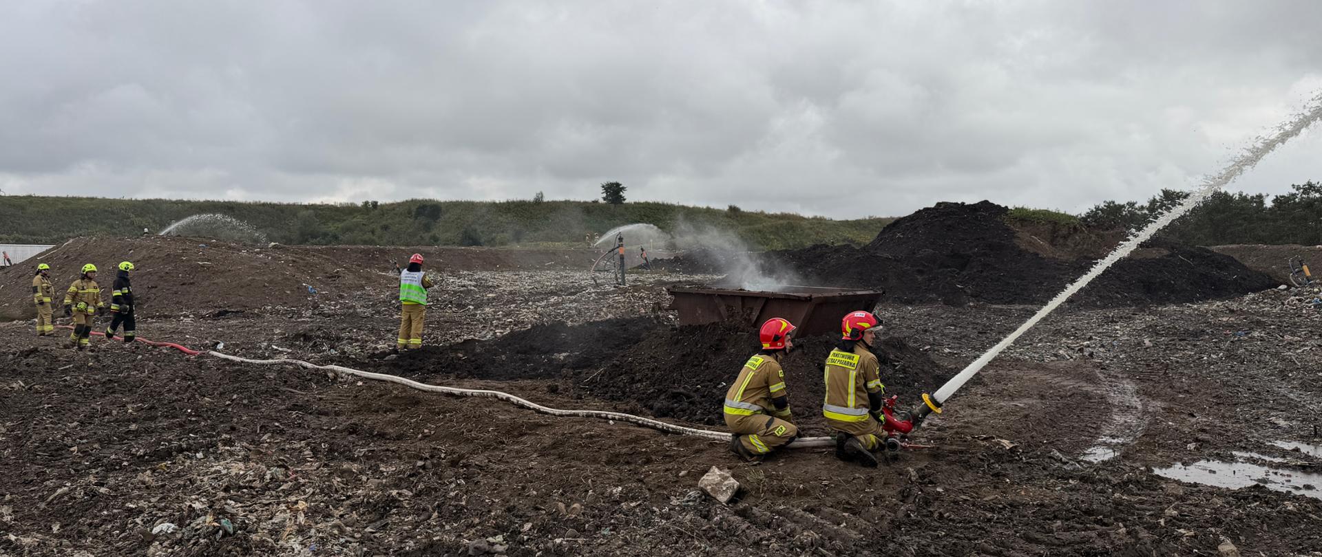 Na tym zdjęciu widać działania ratownicze na terenie składowiska odpadów lub pożaru wysypiska. Na pierwszym planie, w centralnej części, dwóch strażaków w hełmach i kombinezonach, klęcząc, obsługuje wąż strażacki, kierując silny strumień wody na prawo od kadru.
W tle, na rozległym, błotnistym i ciemnym terenie zmieszanym ze śmieciami, widać grupę innych strażaków oraz pojedynczą postać w odblaskowej kamizelce, prawdopodobnie dowódcę akcji. W centralnej części widać dym lub parę unoszącą się z jakiegoś zagłębienia lub kontenera. W oddali widoczne są kolejne strumienie wody na wzgórzu lub hałdzie. Niebo jest pochmurne i szare, co nadaje scenie poważny charakter.
