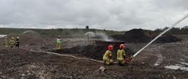 Na tym zdjęciu widać działania ratownicze na terenie składowiska odpadów lub pożaru wysypiska. Na pierwszym planie, w centralnej części, dwóch strażaków w hełmach i kombinezonach, klęcząc, obsługuje wąż strażacki, kierując silny strumień wody na prawo od kadru.
