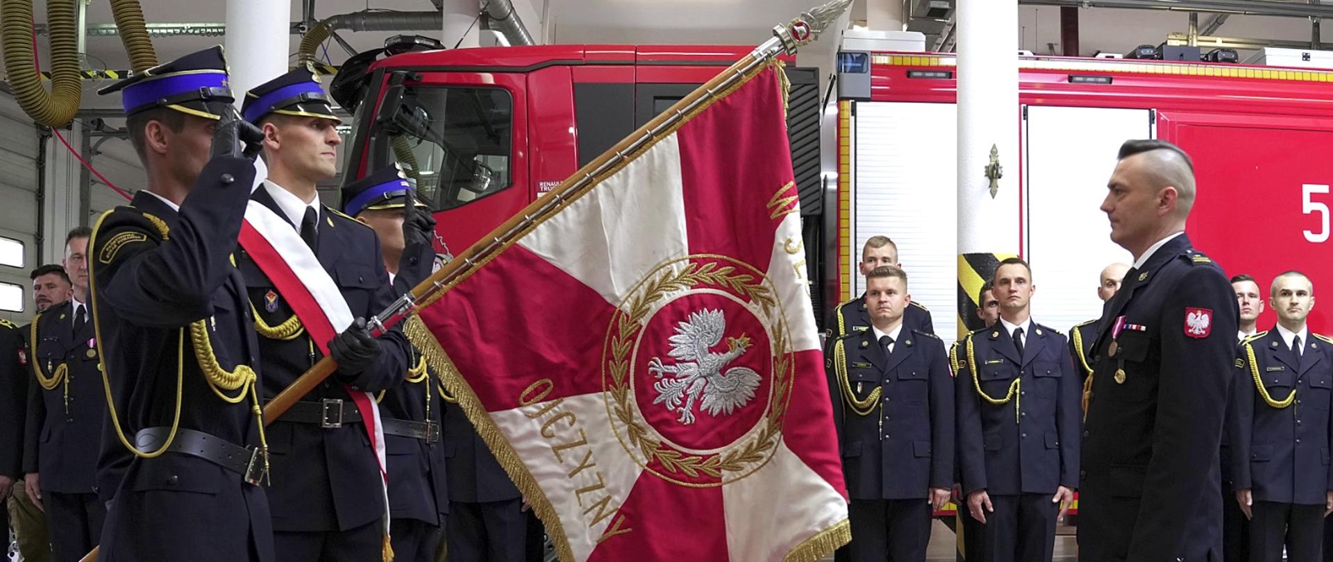 Poczet sztandarowy strażaków w galowych mundurach salutuje przed oficerem podczas uroczystości w komendzie. W tle wóz strażacki i szeregi funkcjonariuszy.
