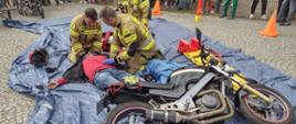 Na zdjęciu widać strażaków udzielających pomocy medycznej nieprzytomnemu motocykliście podczas pokazu z ratownictwa drogowego. Trzech strażaków przeprowadza badanie urazowe. Motocyklista leży na plecach. Obok niego leży motocykl. W tle widać ludzi oglądających pokaz.