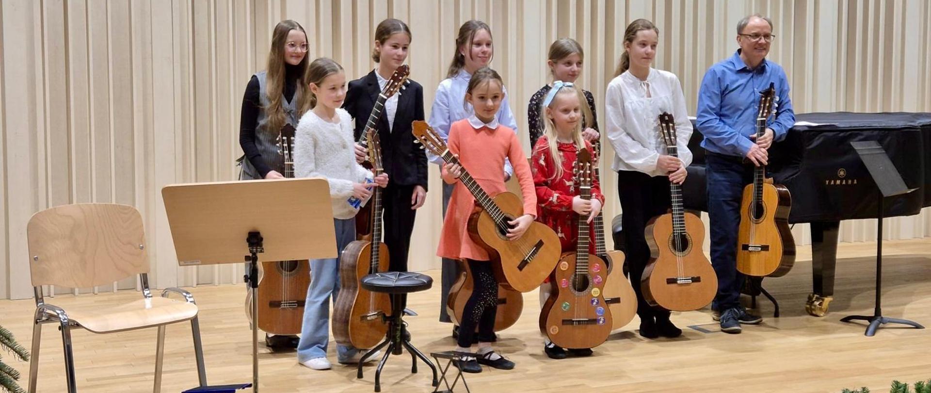 Grupa dzieci i młodzieży stojąca na scenie sali koncertowej, trzymająca gitary klasyczne. Obok nich stoi nauczyciel z gitarą. W tle widoczny fortepian oraz jasna, drewniana ściana akustyczna. Uczestnicy są odświętnie ubrani i ustawieni do pamiątkowego zdjęcia po koncercie.