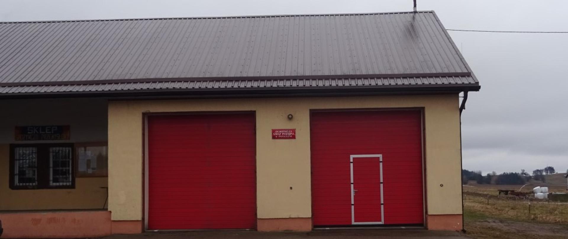 Widoczny budynek koloru żółtego z dwiema bramami koloru czerwonego. Dach w kolorze brązowym, na dachu widoczna syrena alarmowa. Podjazd przed garażami wylany asfaltem. 