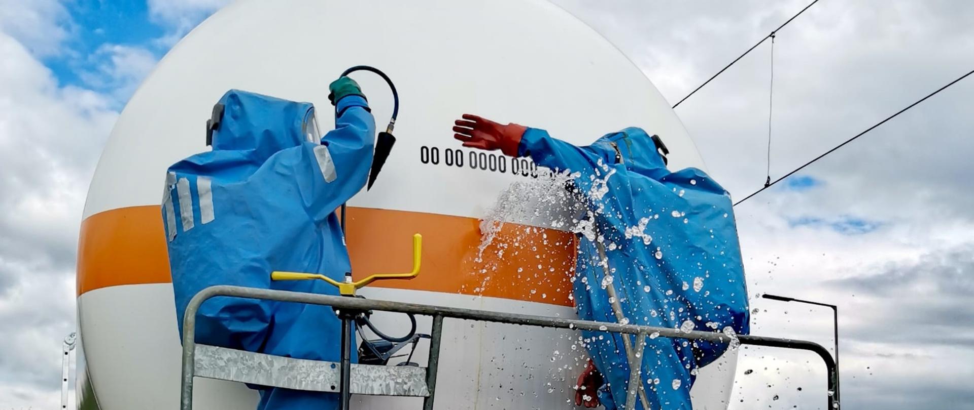 Dwóch strażaków ubranych w gazoszczelne ubrania (zwane CUGAMI) stojących na rampie cysterny kolejowej przystępuje do ograniczenia wycieku substancji z cysterny poprzez użycie korka uszczelniającego