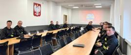 Na zdjęciu widzimy strażaków w czarnych mundurach siedzących w sali wykładowej podczas zajęć teoretycznych. Na ścianie widzimy godło RP Orła Białego w koronie złotej, na drugiej ścianie wyświetlany jest napis Komenda Wojewódzka Państwowej Straży Pożarnej w Kielcach oraz logo PSP.