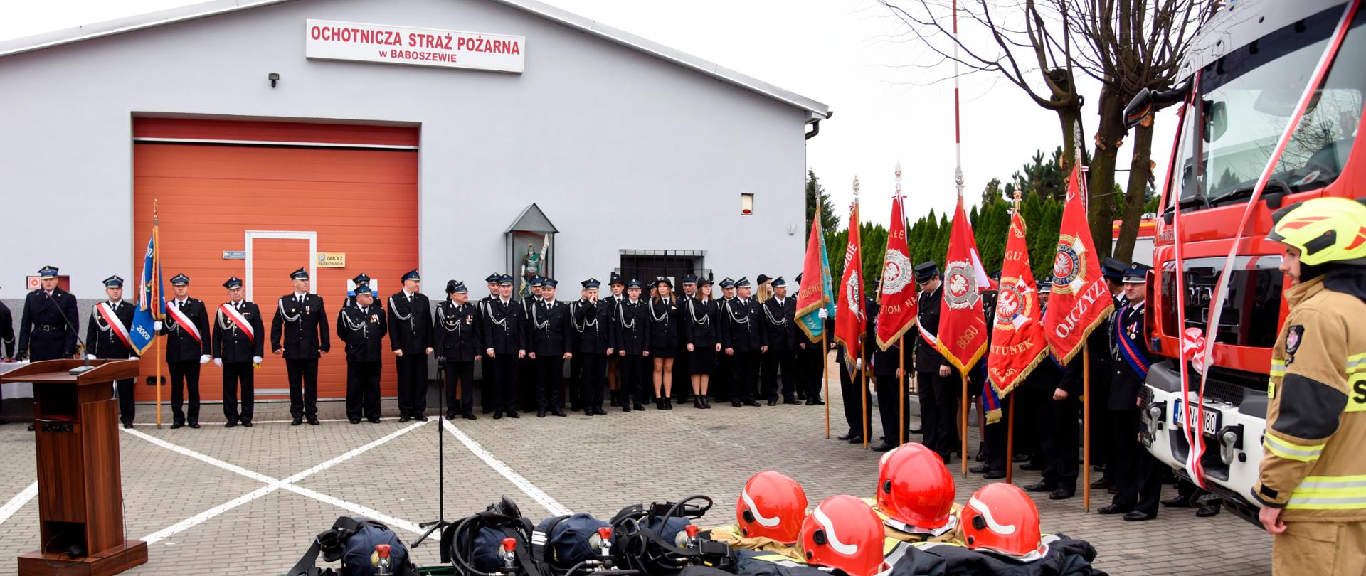 Na pierwszym planie stoi stół na którym znajdują się: sprzęt pożarniczy oraz hełmy i ubrania strażackie. W tle stoją strażacy przed budynkiem strażnicy OSP.