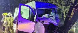 Na zdjęciu biały bus dostawczy po uderzenie w przy drożne drzewo. Jest noc, występuje oświetlenie sztuczne z pojazdów ratowniczych. W tle działający strażacy ubrani w ubrania specjalne.