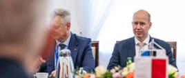 Na zdjęciu widać ministra Igora Taro (po prawej) oraz innego członka delegacji estońskiej – obydwaj siedzą przodem do obiektywu. Po lewej stronie, siedzący tyłem do obiektywu, minister Tomasz Siemoniak. Po prawej stronie widać proporczyki – polski (biało-czerwony) i estoński (niebiesko-czarno-biały). Na stole: termosy z kawą, filiżanki, szklanki, butelki z wodą, talerzyki z przekąskami oraz kompozycja kwiatowa. W tle okno z białą zasłoną.