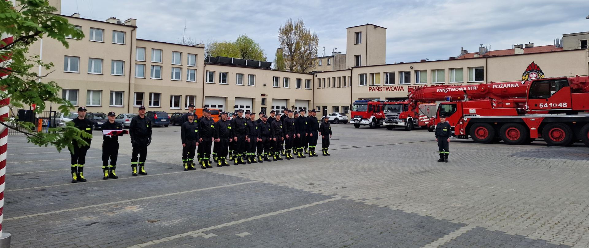 Na zdjęciu znajdują się funkcjonariusze Państwowej Straży Pożarnej w Tczewie. Poczet flagowy podczas, pododdział składający się z dwóch zmian służbowych strażaków JRG Tczew. Z prawej strony Widoczny dowódca uroczystości. W tle stoją samochody gaśnicze i specjalne z JRG Tczew.