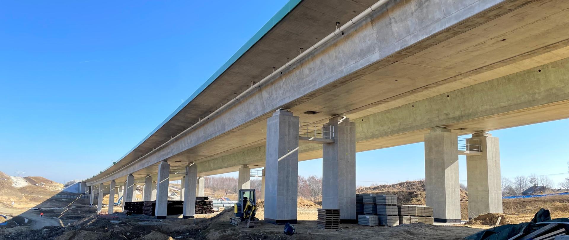Świeci słońce. Widok z dołu, z poziomu ziemi na budowaną estakadę. Dookoła obiektu widać sprzęt budowlany, złożone materiały budowlane.