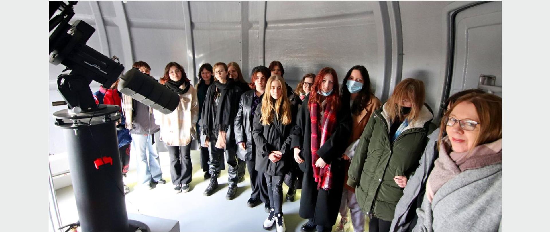 Grupa młodzieży w pomieszczeniu obserwatorium stojąca przed lunetą.