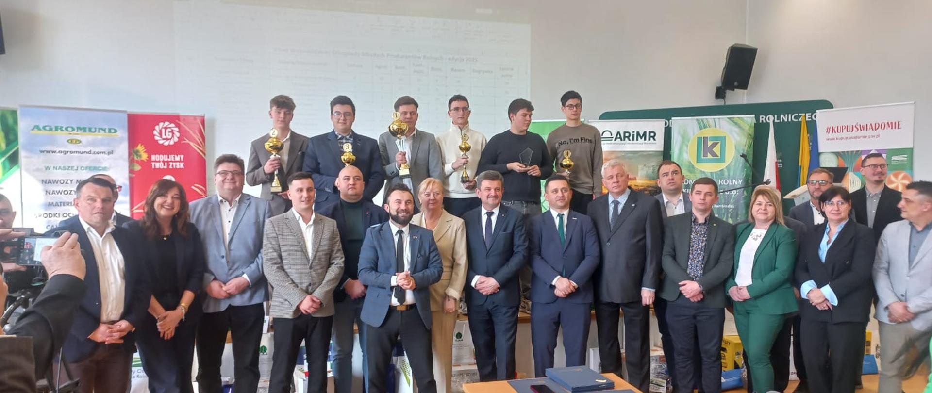 Duża grupa osób pozuje do zdjęcia w sali z plakatami i rollupami instytucji rolniczych. Sześciu młodych mężczyzn stoi z tyłu na podwyższeniu i trzyma w rękach puchary.