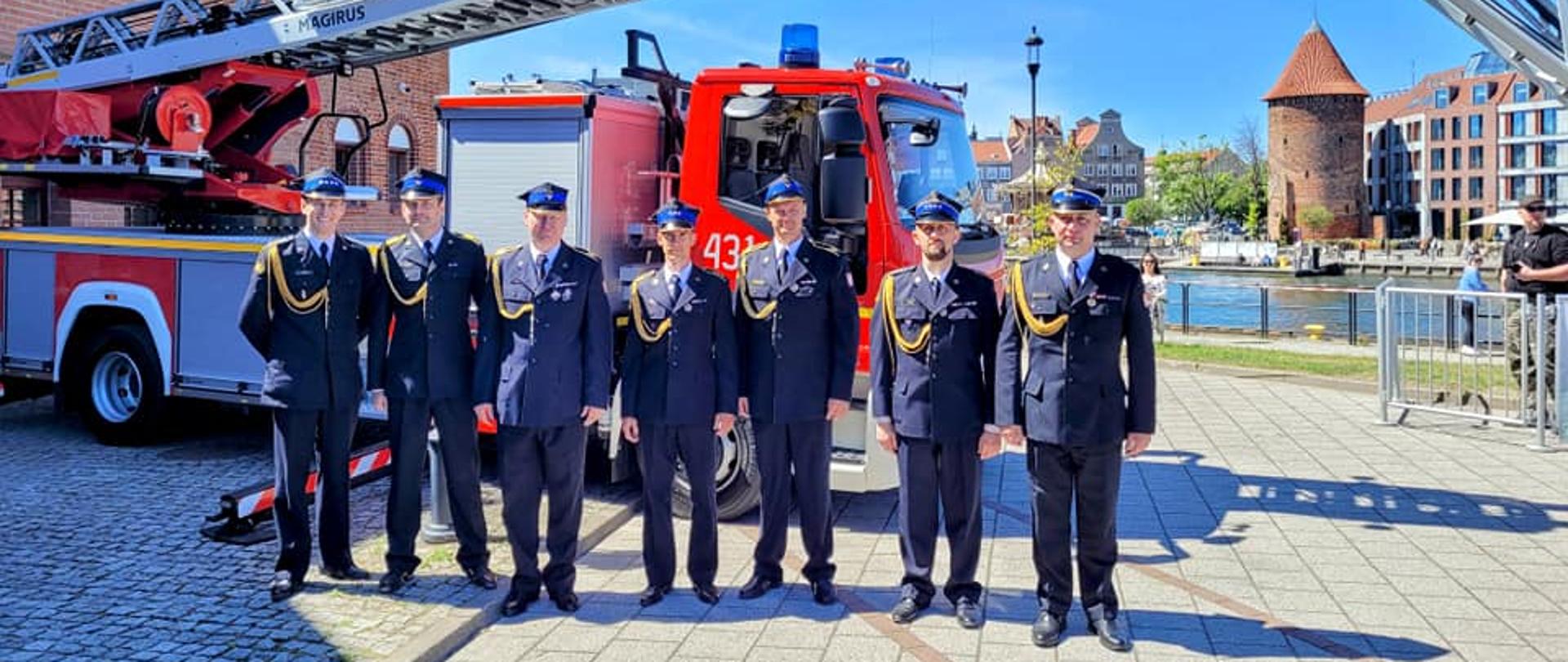 Strażacy w mundurach galowych na tle podnośnika. W oddali stare miasto w Gdańsku.