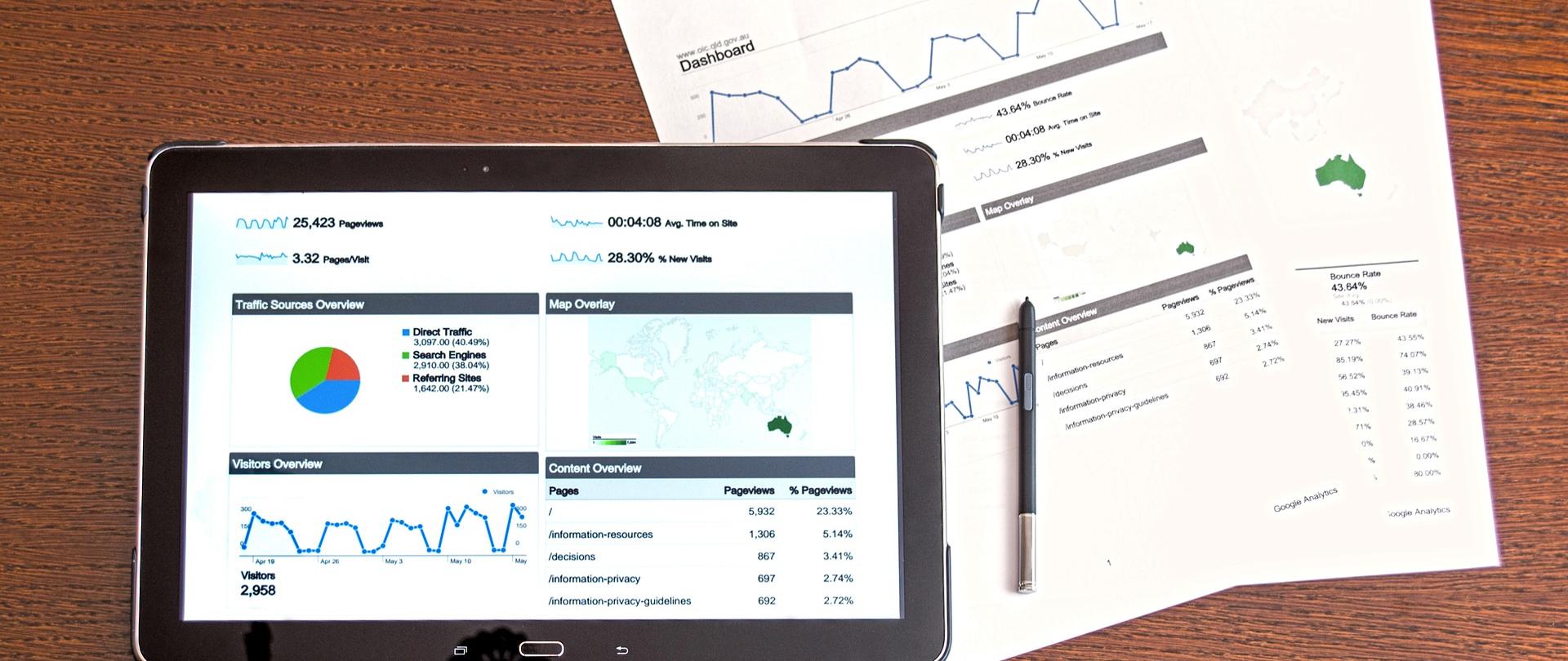 Na zdjęciu znajduje się tablet z plikami z różnymi wykresami. Obok tabletu leżą kartki z danymi statystycznymi, a na nich leży długopis.