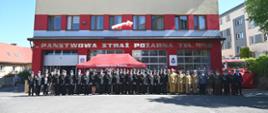 Kolorowa fotografia wykonana pogodny dzień na zewnątrz. Przedstawia strażaków PSP i OSP oraz policjantów podczas uroczystości z okazji Dnia Strażaka. Strażacy stoją na tle garaży Komendy PSP w Brzozowie w ubraniach wyjściowych i służbowych.