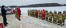 Strażacy PSP i OSP ustawieni w szeregu na brzegu zamarzniętego jeziora podczas odprawy przed rozpoczęciem ćwiczeń z ratownictwa lodowego. W tle widoczny akwen pokryty lodem oraz pojazd służbowy.